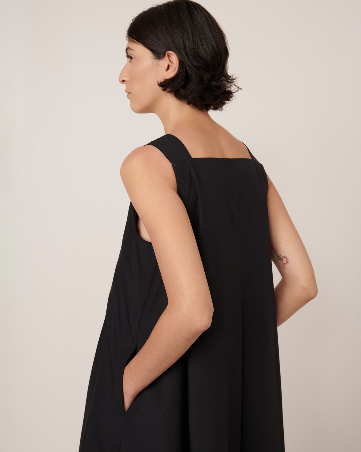A woman with short dark hair is wearing the Kowtow Study Dress in black—a sleeveless, standard-fit dress—standing with her hands in her pockets and facing left.