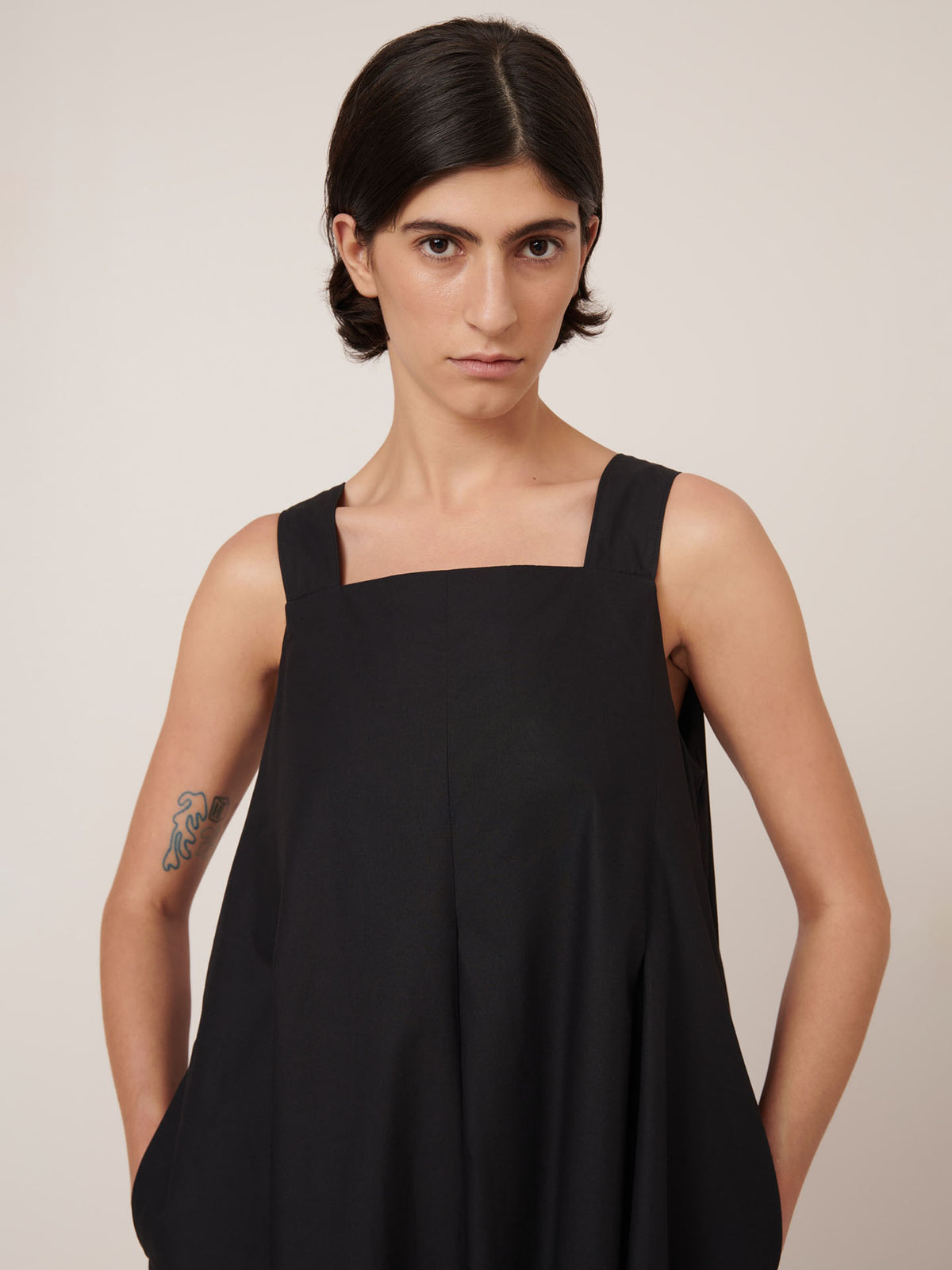 A person with short dark hair wears the Kowtow Study Dress in black—a standard fit, sleeveless style—standing hands in pockets and facing the camera against a plain background.