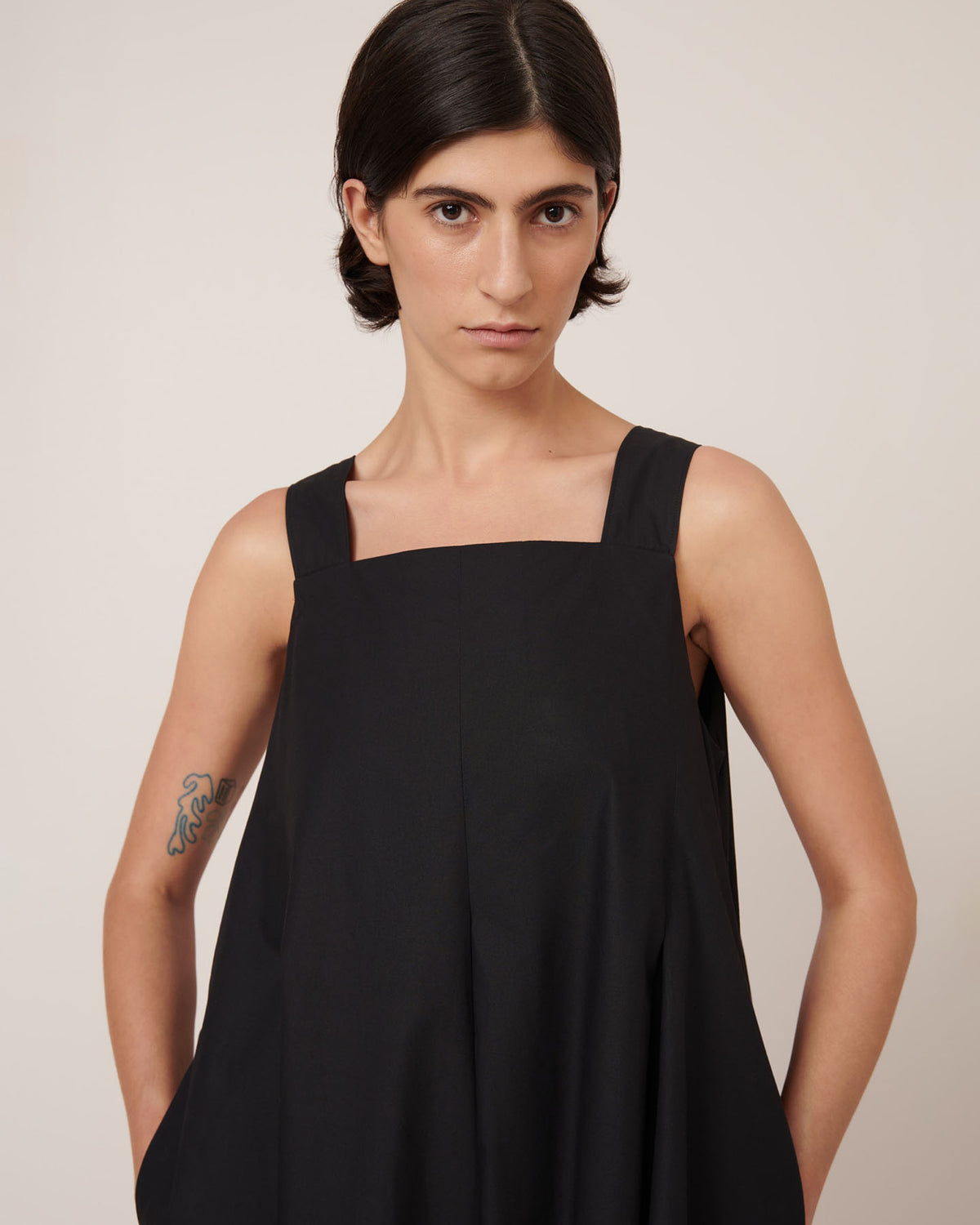 A person with short dark hair wears the Kowtow Study Dress in black—a standard fit, sleeveless style—standing hands in pockets and facing the camera against a plain background.