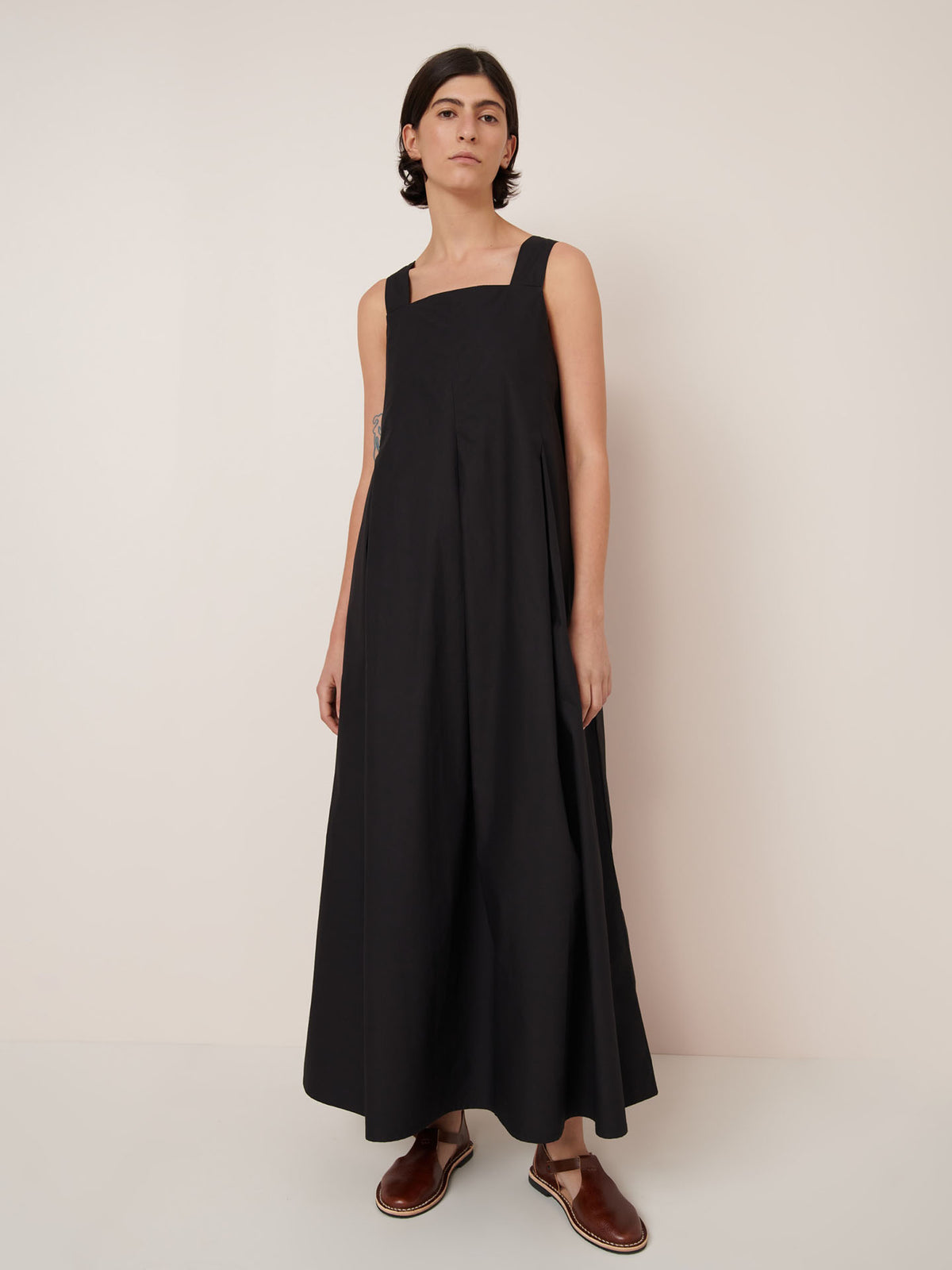 A person stands against a plain background wearing the Kowtow Study Dress – Black, a sleeveless, ankle-length style paired with brown sandals. Showcasing the standard fit—see our size guide for measurements.