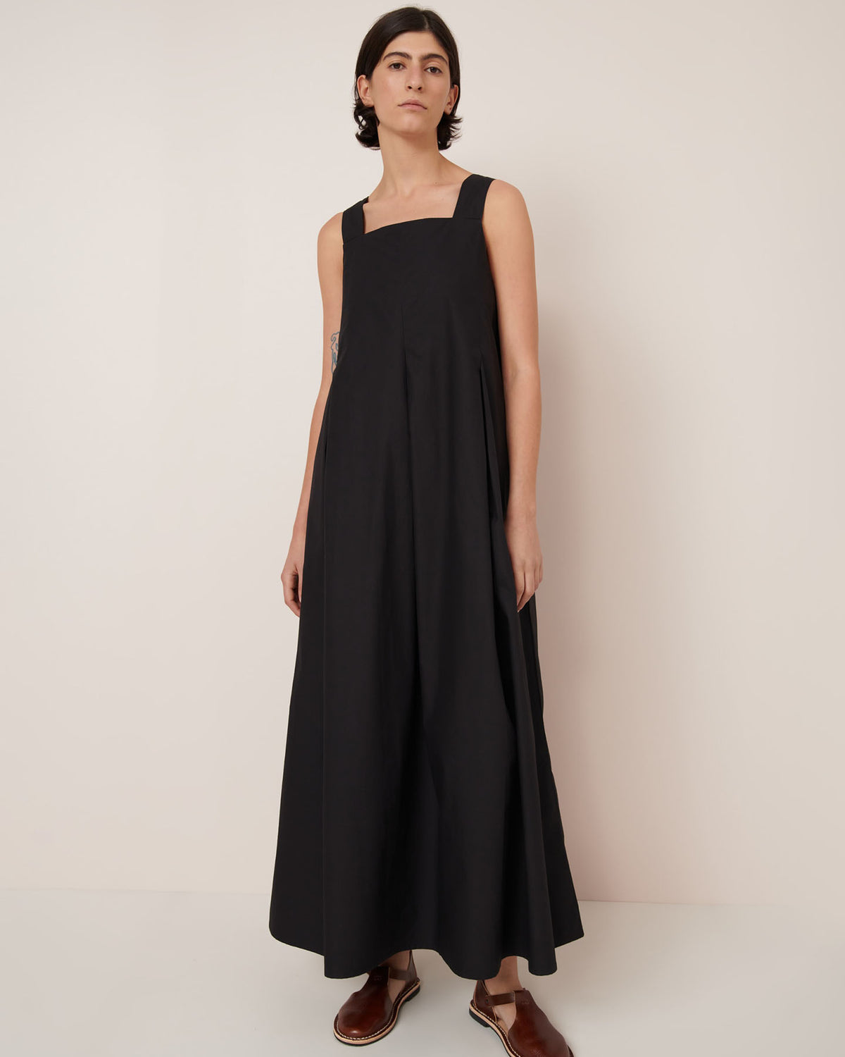 A person stands against a plain background wearing the Kowtow Study Dress – Black, a sleeveless, ankle-length style paired with brown sandals. Showcasing the standard fit—see our size guide for measurements.