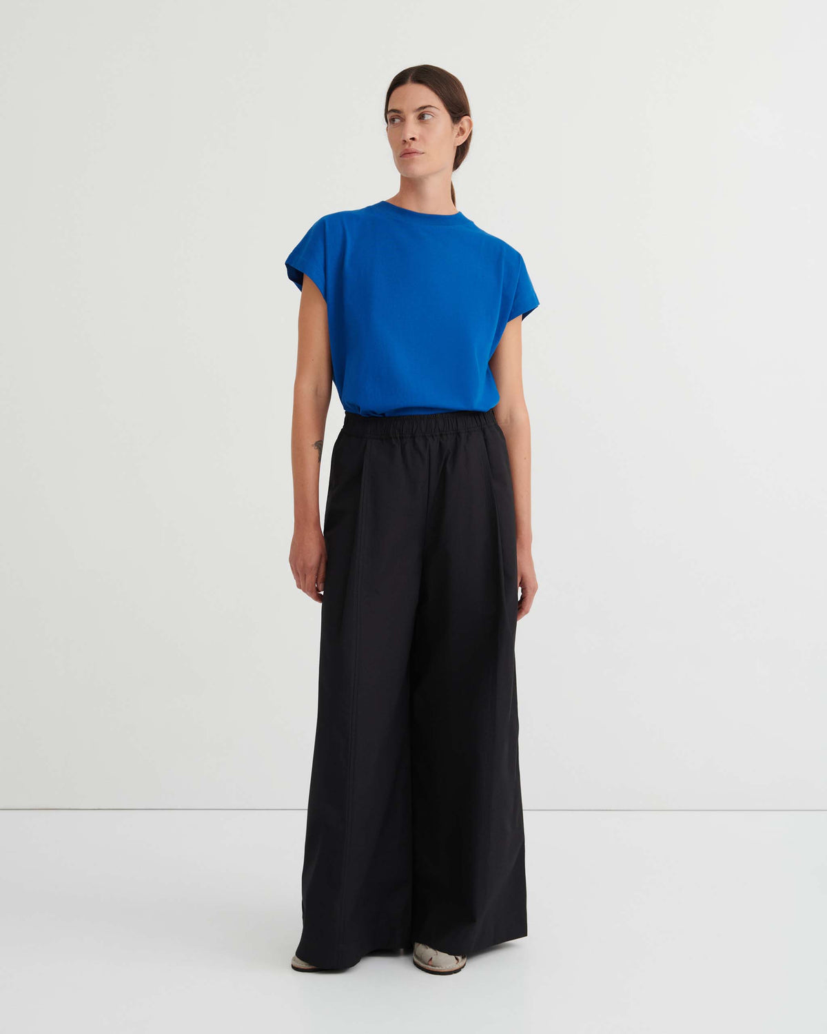 Person standing in a plain room, wearing a blue short-sleeve top, wide-leg black pants, and white shoes.