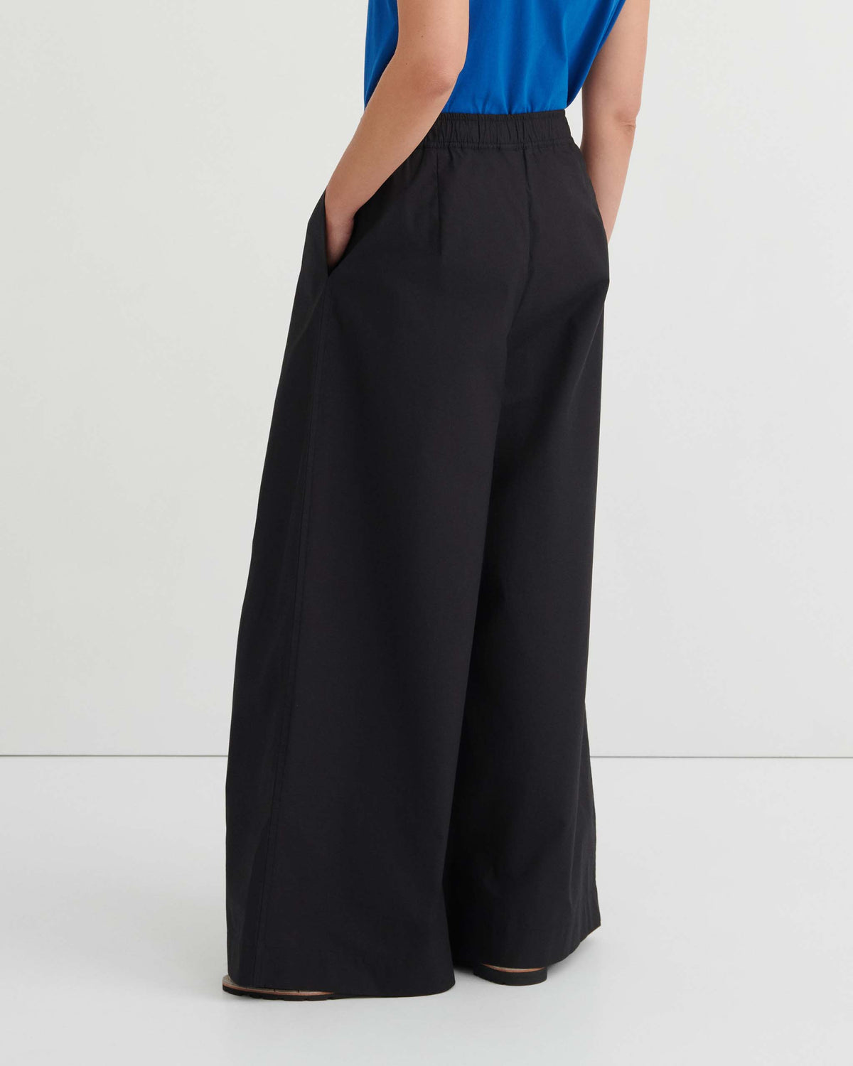 A person stands against a white wall wearing wide-leg black pants and a blue top, with hands in their pockets.