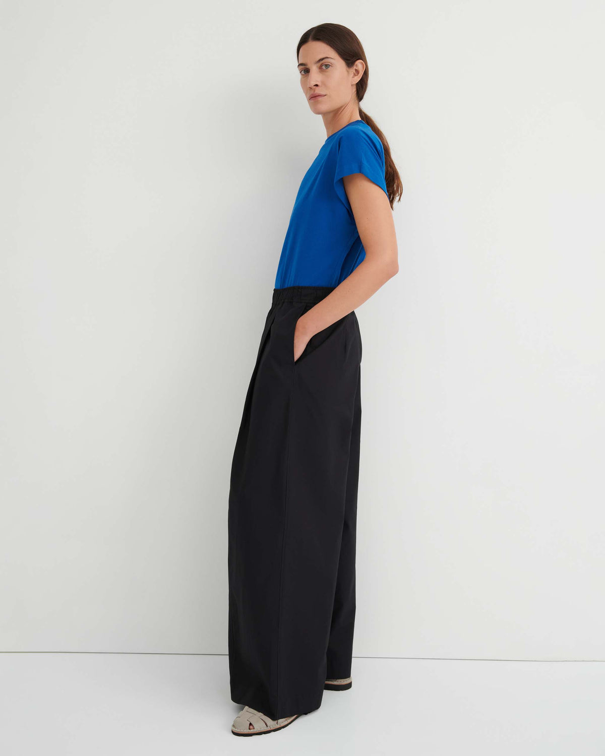 Person standing sideways against a white wall, wearing a blue t-shirt, black wide-leg pants, and beige shoes, with hands in pockets and long hair tied back.
