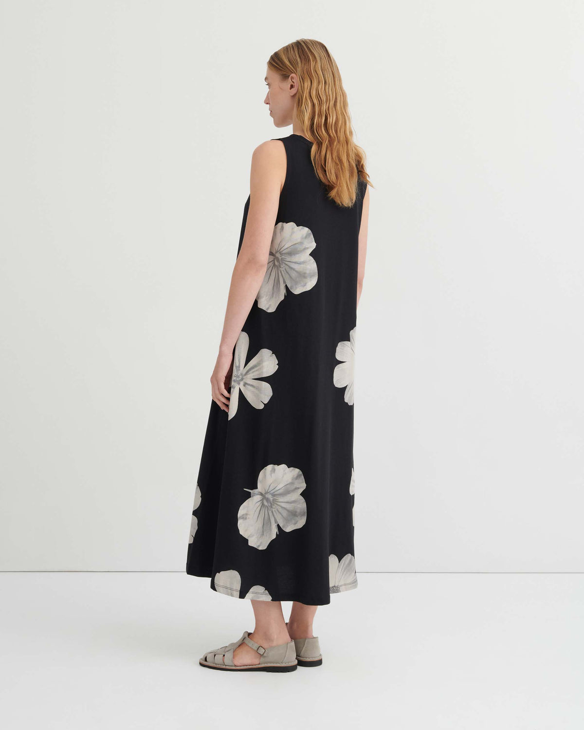 A woman in profile wearing a black sleeveless maxi dress with a large white floral print and gray sandals.