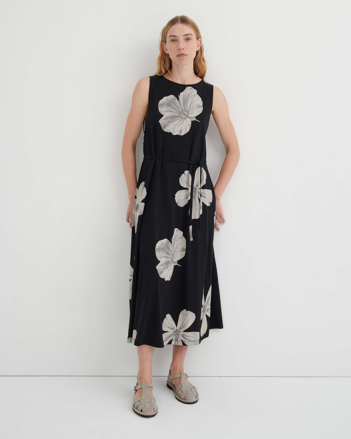 A woman models a black sleeveless midi dress with a white floral print and a tie waist, paired with grey closed-toe sandals.