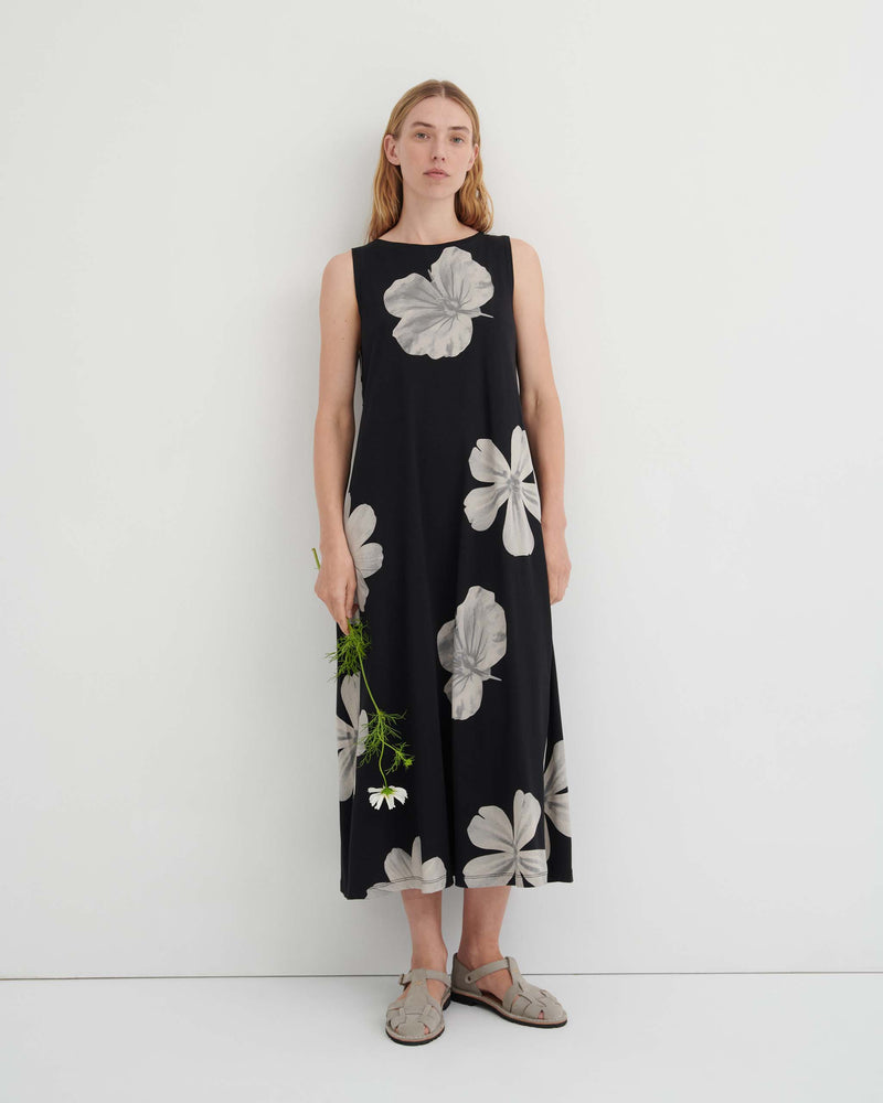 A woman wearing a black sleeveless midi dress with a large grey floral pattern and grey strappy sandals, holding a single white flower.