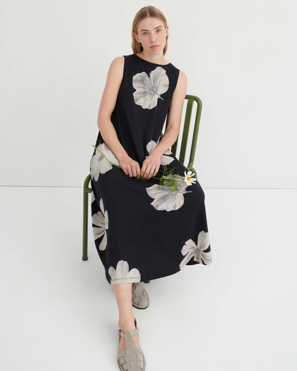 A person wearing a black sleeveless dress with a white floral print sits on a green chair holding a daisy.