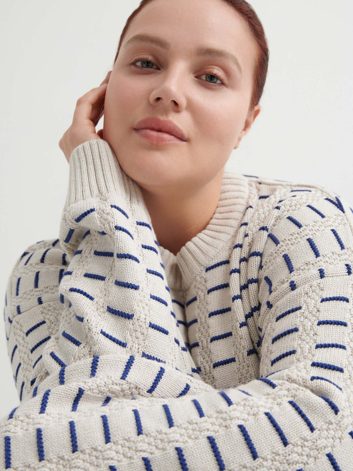 A person with fair skin and brown hair wears a Kowtow Thread Jumper – Oat Melange in a relaxed fit, resting their head on their hand and looking at the camera.