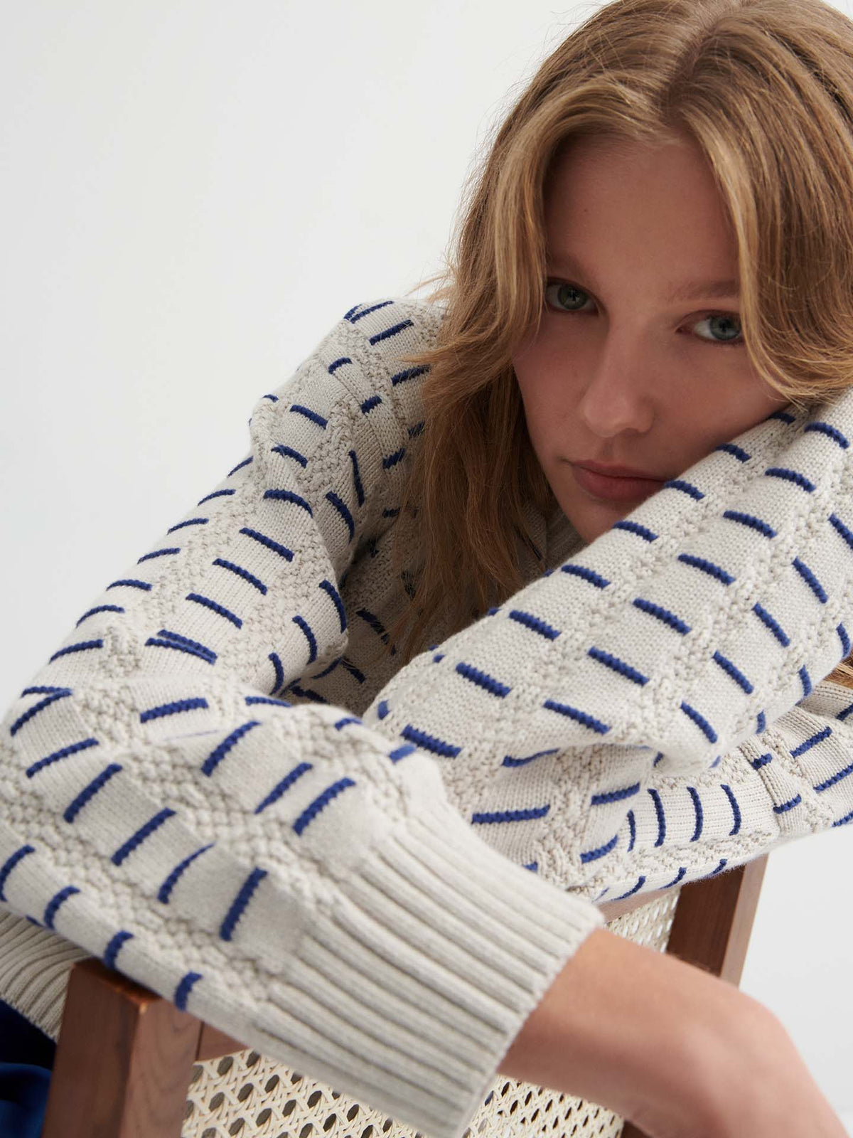 A person with long, light brown hair leans forward on a wooden chair, wearing the Thread Jumper – Oat Melange in a relaxed cream sweater with blue and navy stripes, and looks directly at the camera.