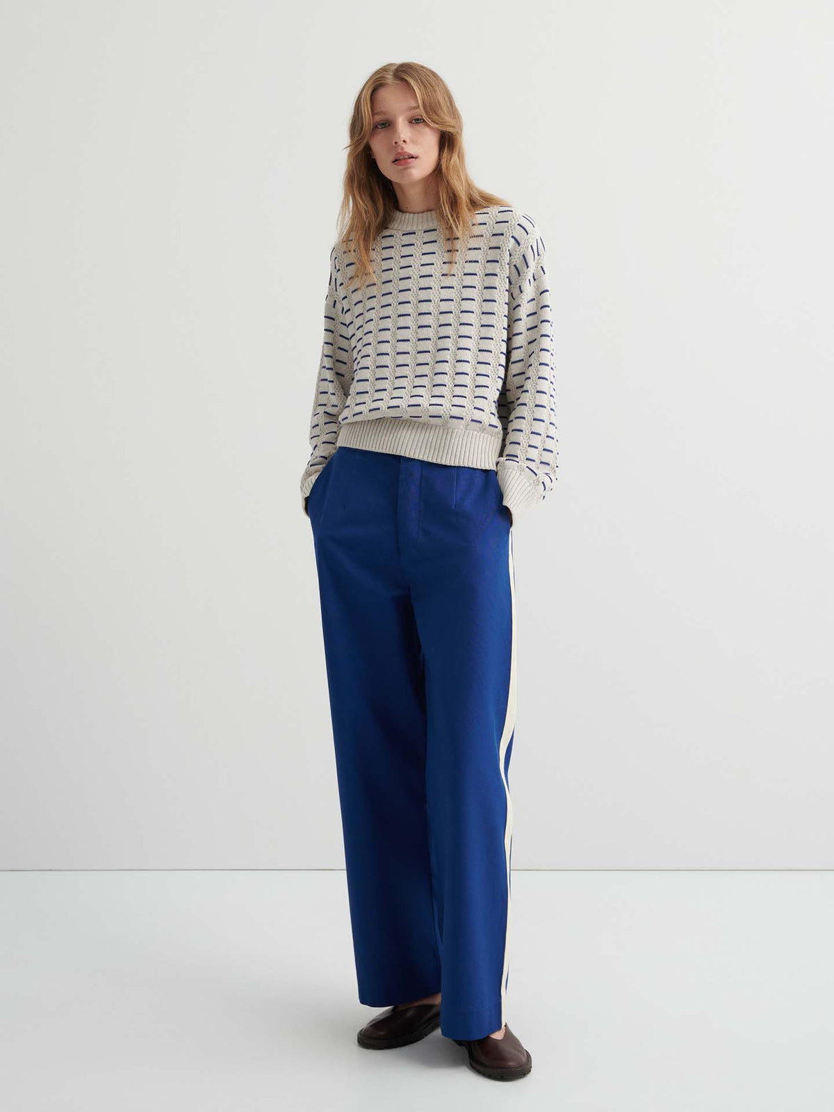 A woman stands against a plain background wearing the Thread Jumper – Oat Melange with a relaxed fit, wide-leg blue pants with white side stripes, and dark shoes.