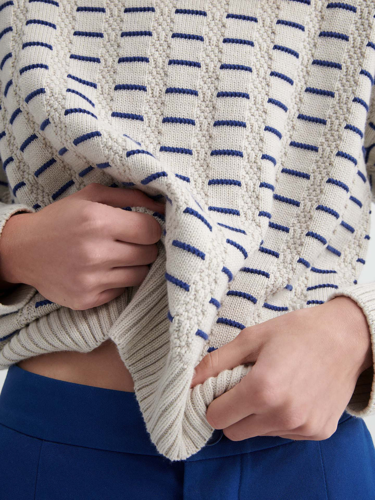 A person in blue pants lifts a relaxed fit cream-colored Thread Jumper – Oat Melange with blue stripes from Kowtow, revealing part of their midriff.