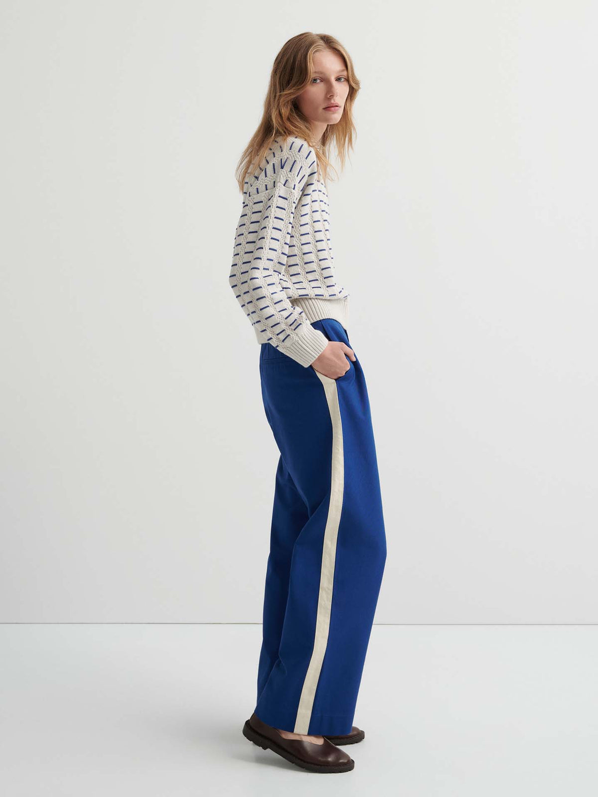 A woman stands sideways wearing a Thread Jumper – Oat Melange, relaxed fit blue wide-leg pants with a white side stripe, and dark shoes against a plain white background.
