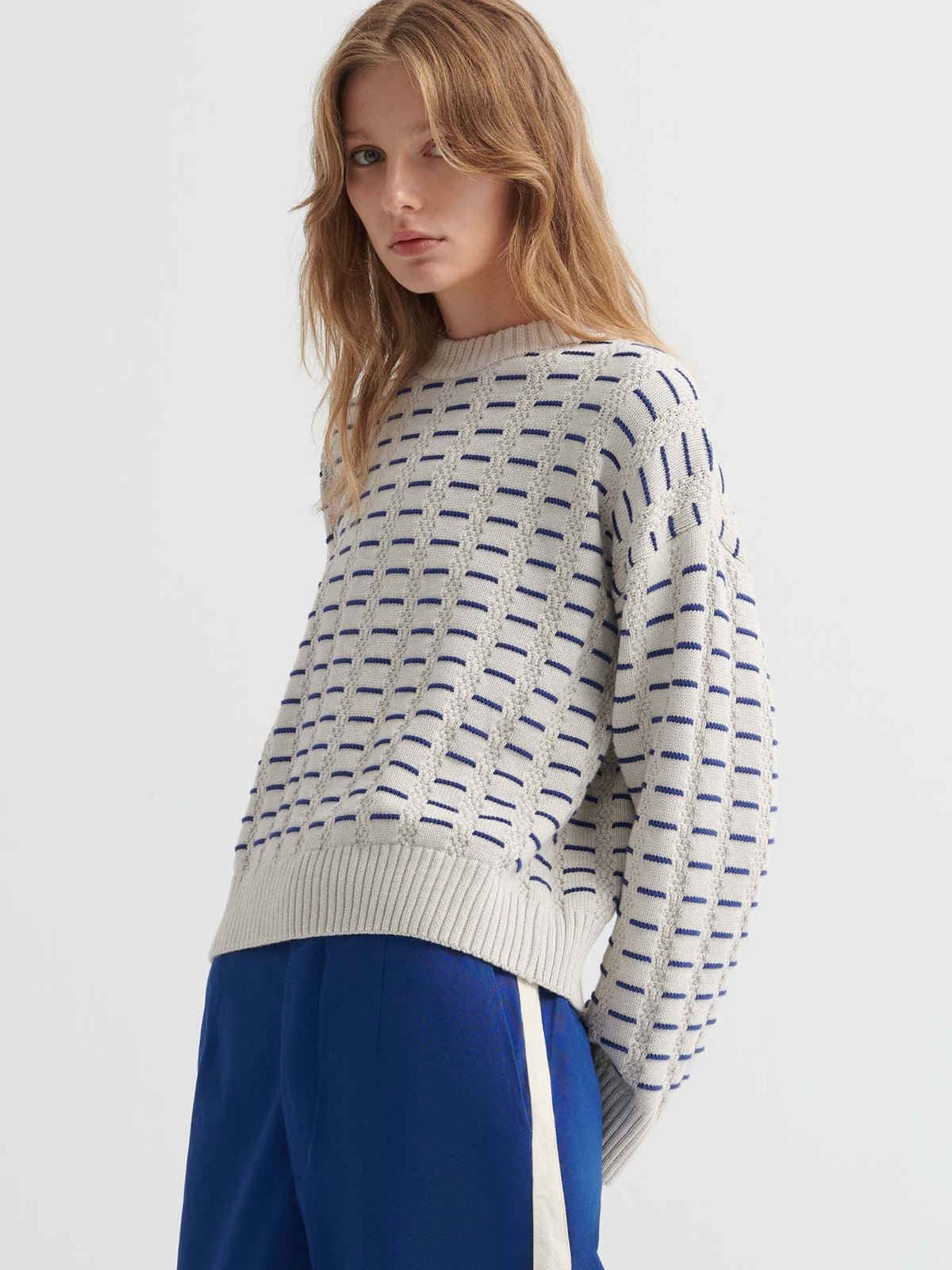 A person wears the Thread Jumper – Oat Melange, a cream and blue textured sweater in a relaxed fit, paired with blue pants with a white side stripe, standing against a plain white background.