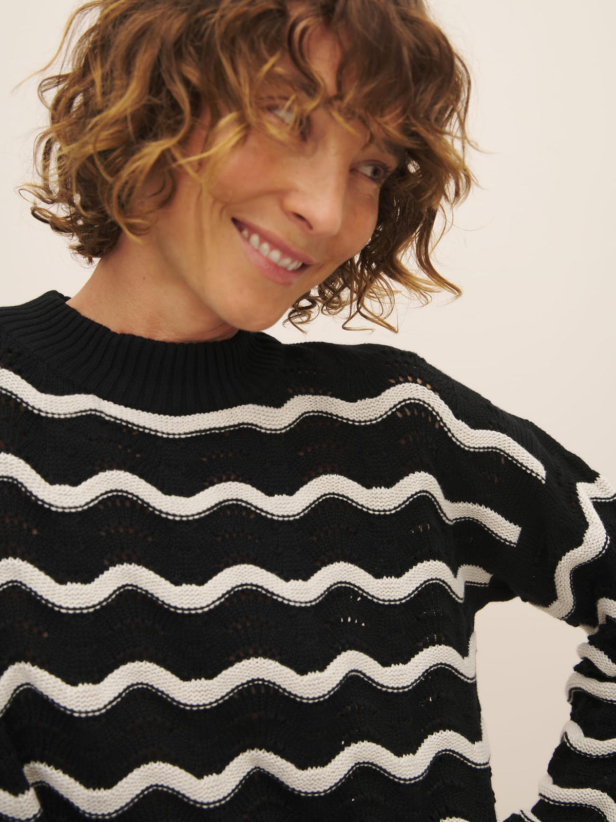 A person with curly hair smiling and wearing a Tide Jumper, a plastic-free black sweater with white wave patterns made from Fairtrade organic cotton by Kowtow.