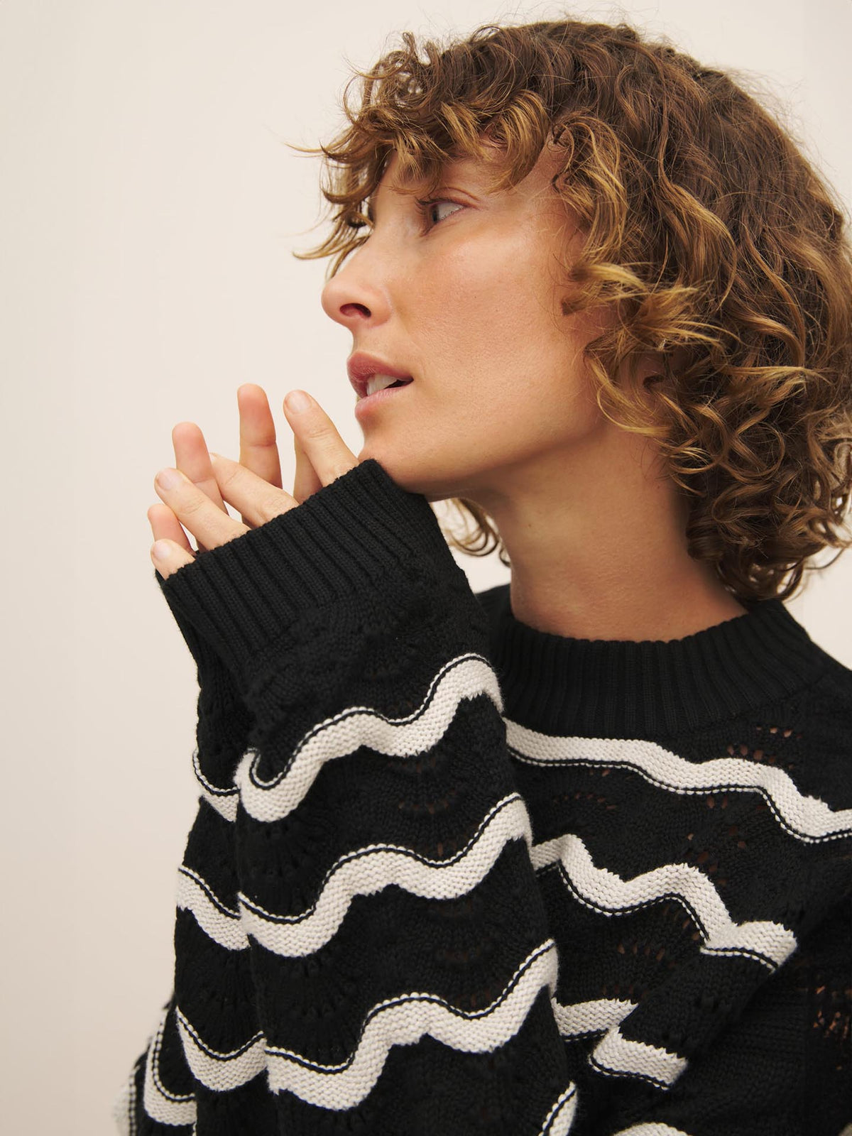 Pensive individual with curly hair wearing a black and white Kowtow wave pattern sweater, gazing to the side.