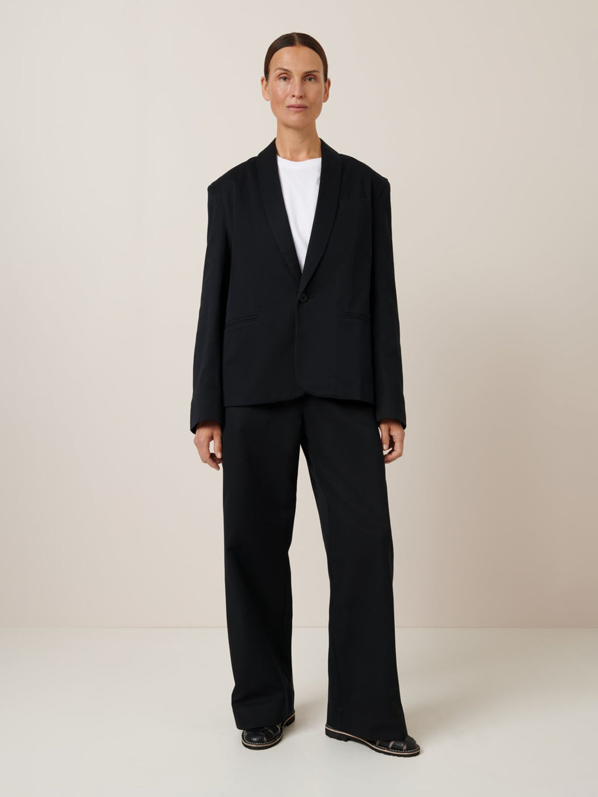 Against a plain backdrop, a person wears an oversized Kowtow Tux Jacket in black with a relaxed fit over a white shirt, their hands resting calmly by their sides.