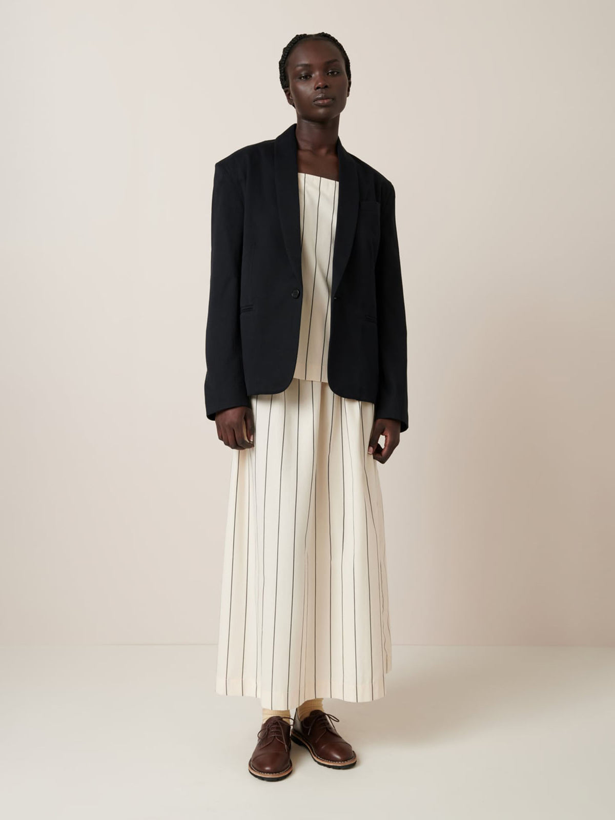 A person in a long, striped cream dress with a relaxed fit pairs it with Kowtow&#39;s Tux Jacket in black and brown shoes, standing against a plain background.