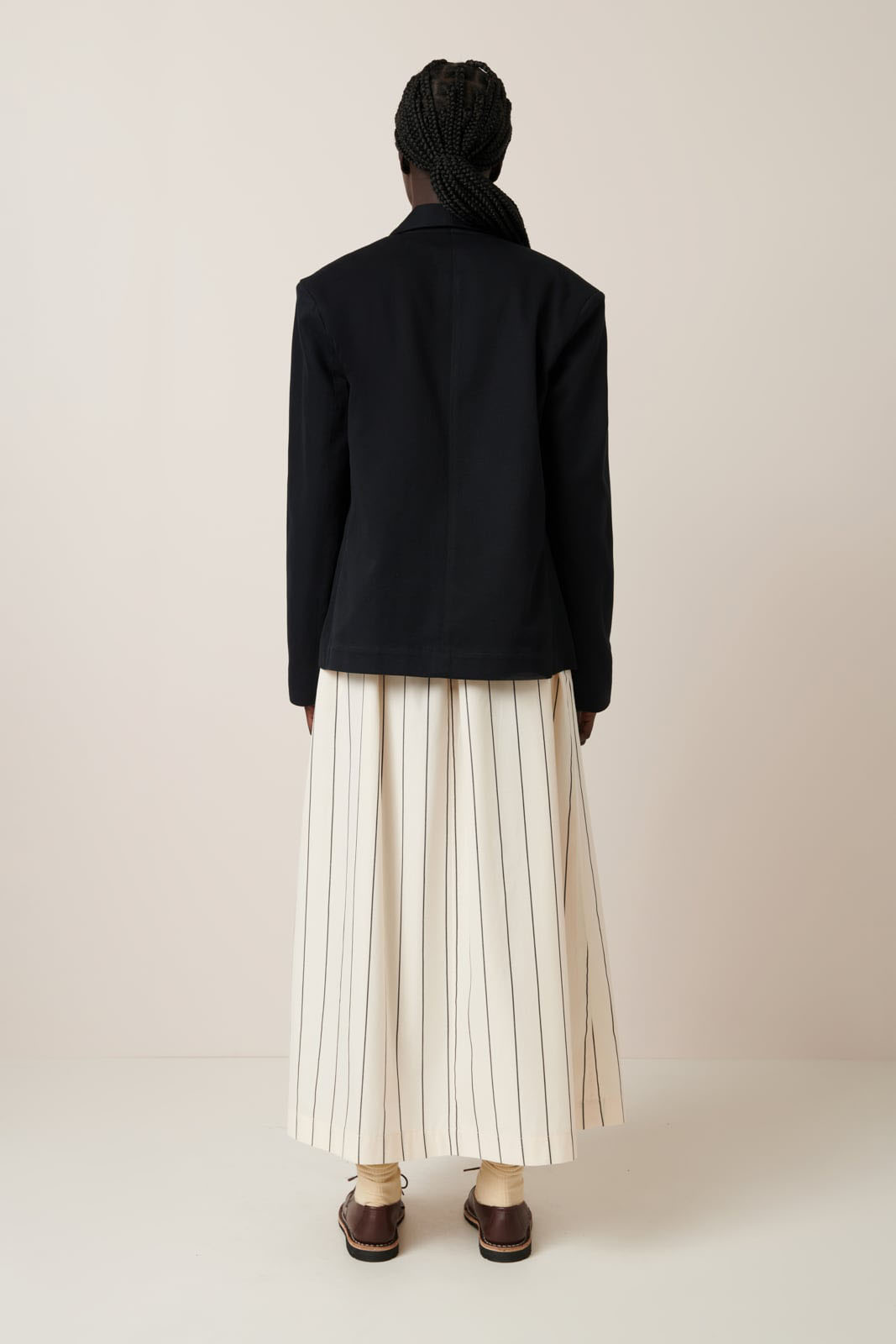 A person with braided hair wearing the Kowtow Tux Jacket in black and a white striped skirt, facing away against a neutral background to showcase the garment pattern.