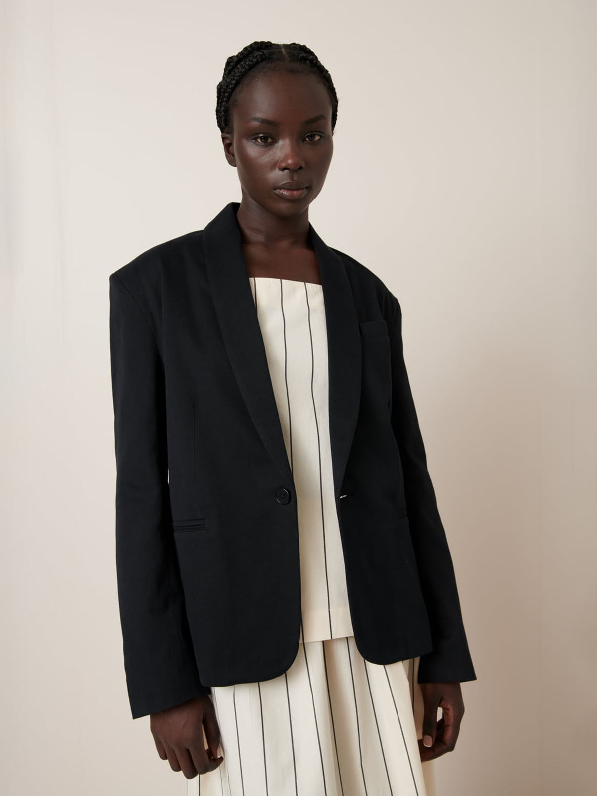 A person poses in a Kowtow Tux Jacket – Black over a white vertically-striped outfit, gazing at the camera.