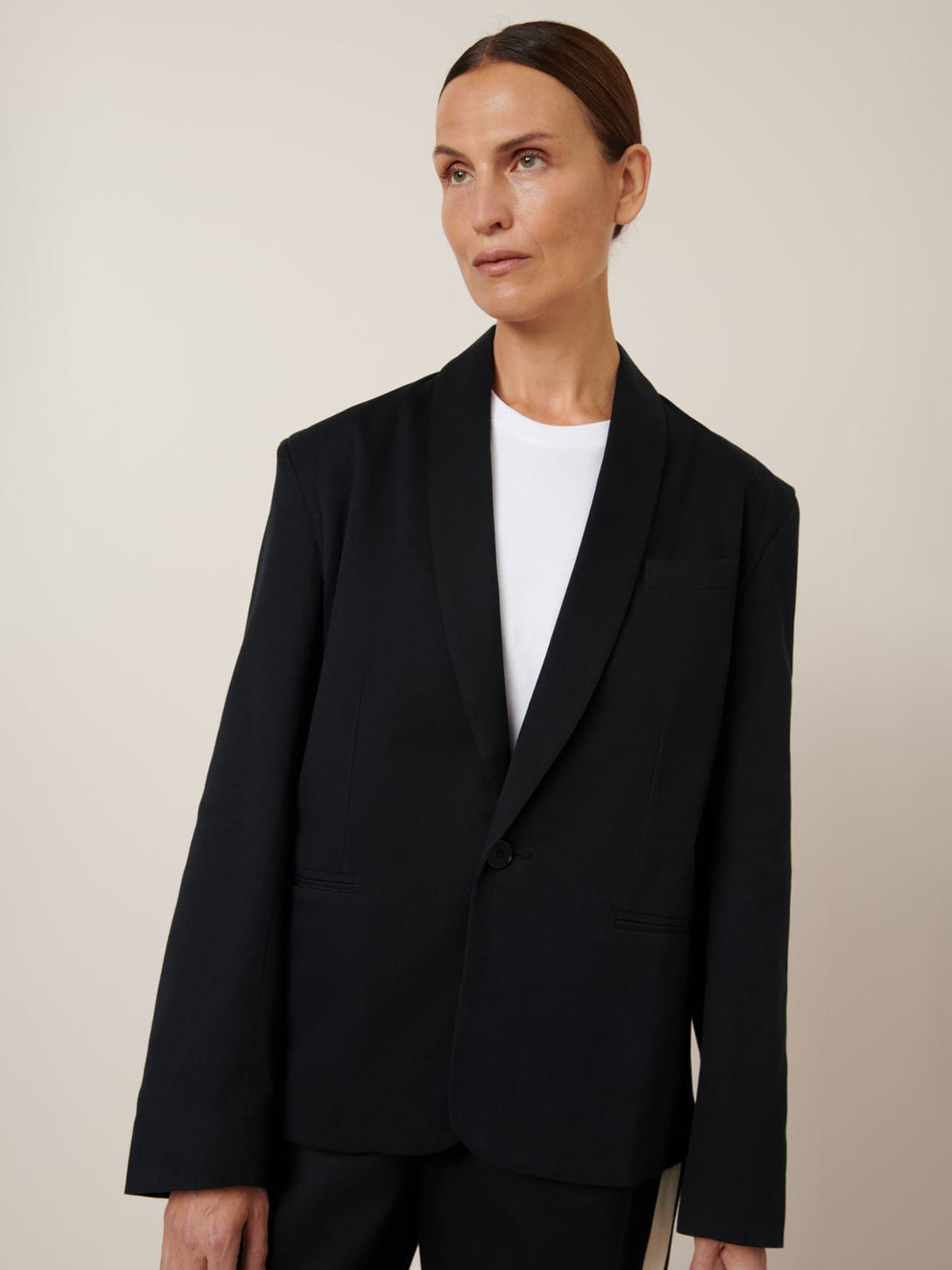 Wearing the Tux Jacket – Black by Kowtow over a white shirt, a person stands against a neutral background, showcasing its relaxed fit that exudes effortless elegance.