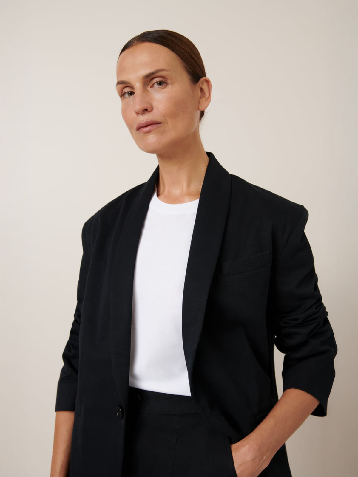 A person wearing the Kowtow Tux Jacket – Black over a white shirt, exhibits a relaxed fit with one hand in their pocket against a plain backdrop.