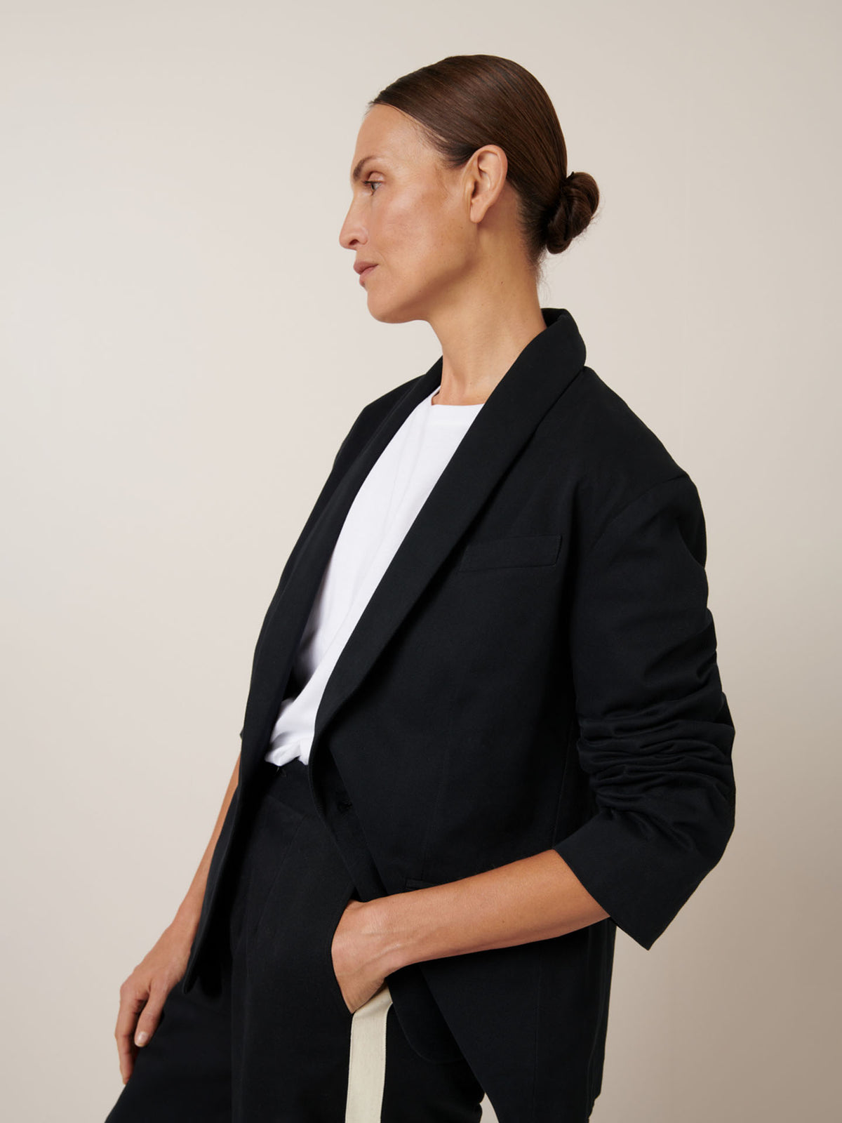 Displaying a relaxed fit, a person poses sideways against a plain backdrop, wearing the Kowtow Tux Jacket in black over a white shirt, with one hand casually in their pocket.