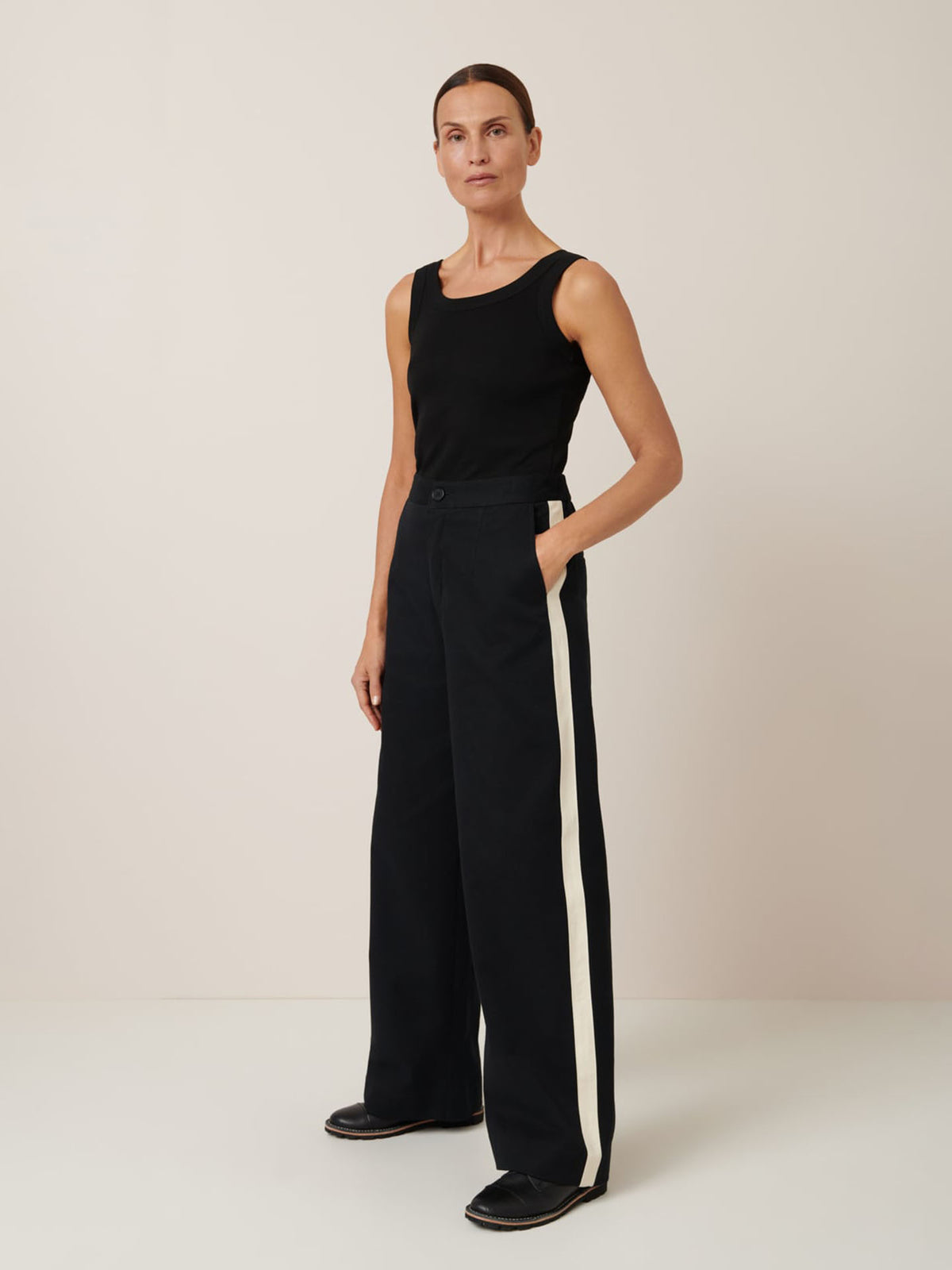 A person models the Kowtow Tux Pant – Black, featuring a trendy mid-rise, relaxed fit, and white side stripes. Styled with a black sleeveless top, they stand against a plain background.