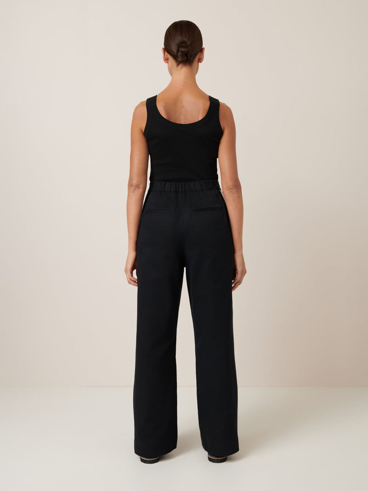 Facing away in a plain room, a person wears the Kowtow Tux Pant – Black paired with a black tank top.