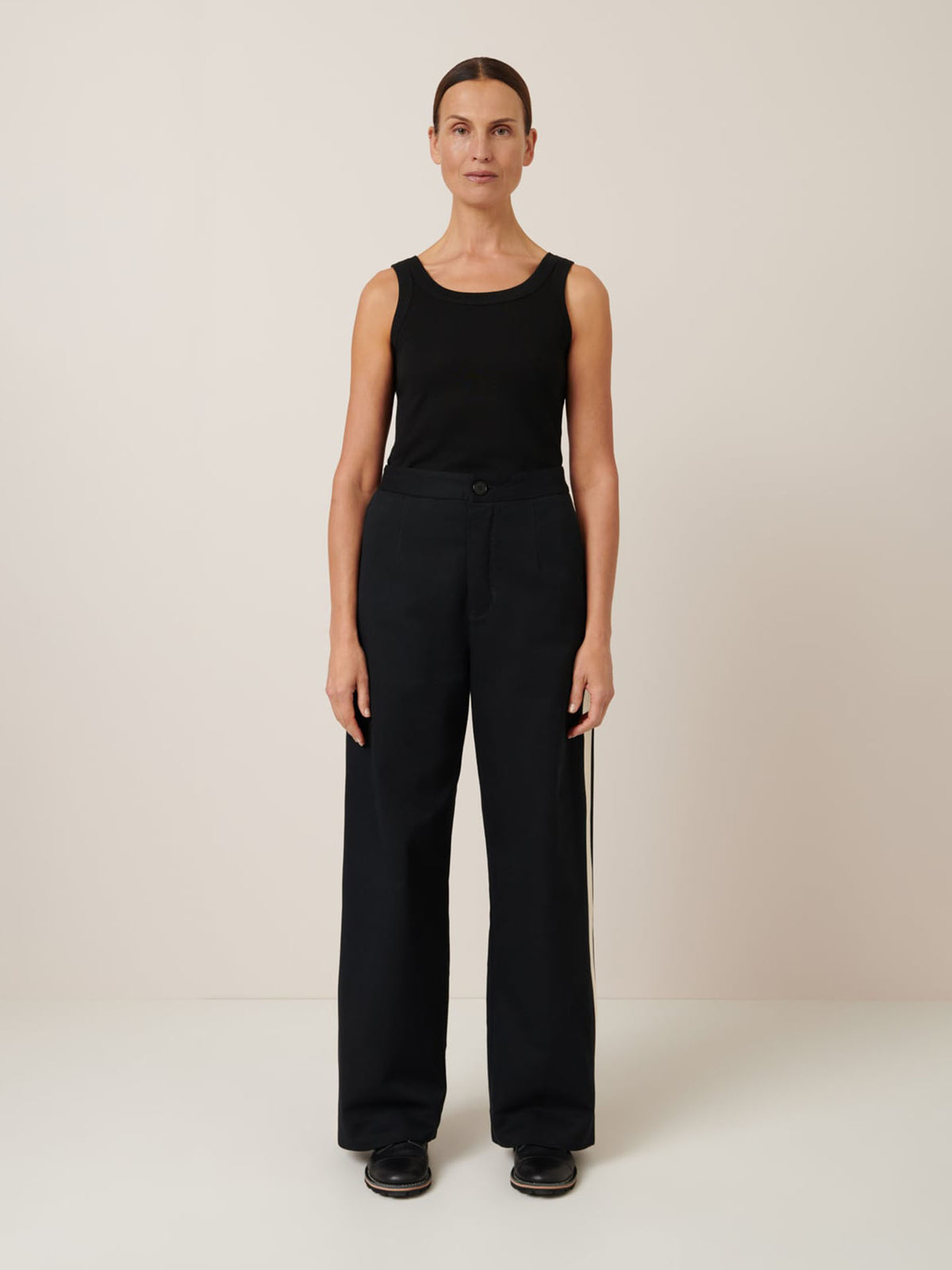 A woman in a black sleeveless top and Kowtow&#39;s Tux Pant – Black, featuring a relaxed-fit and wide-leg design, stands against a plain white background.