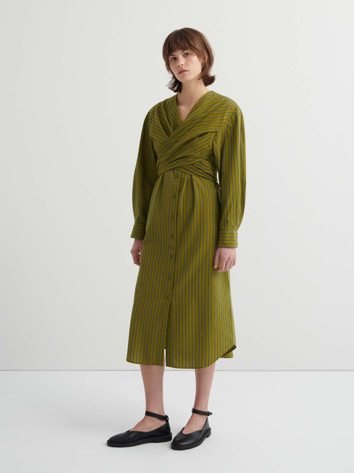 Someone wearing the Twist Dress – Artisan Stripe, a long-sleeve olive green striped dress with a wrapped bodice, button-down front, and relaxed fit, stands against a plain white background.