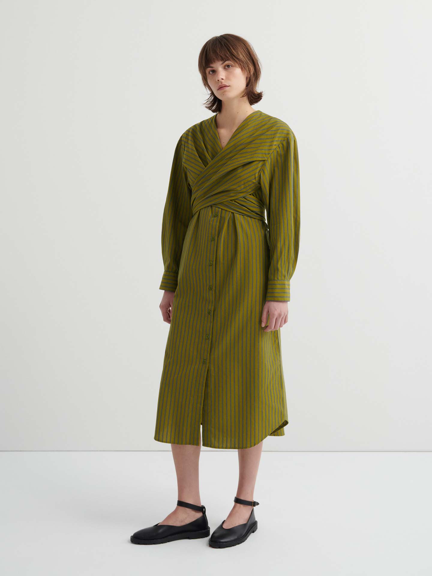 Someone wearing the Twist Dress – Artisan Stripe, a long-sleeve olive green striped dress with a wrapped bodice, button-down front, and relaxed fit, stands against a plain white background.