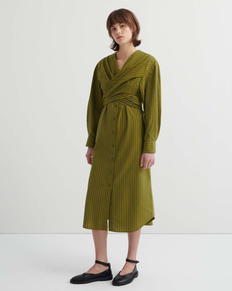 Someone wearing the Twist Dress – Artisan Stripe, a long-sleeve olive green striped dress with a wrapped bodice, button-down front, and relaxed fit, stands against a plain white background.