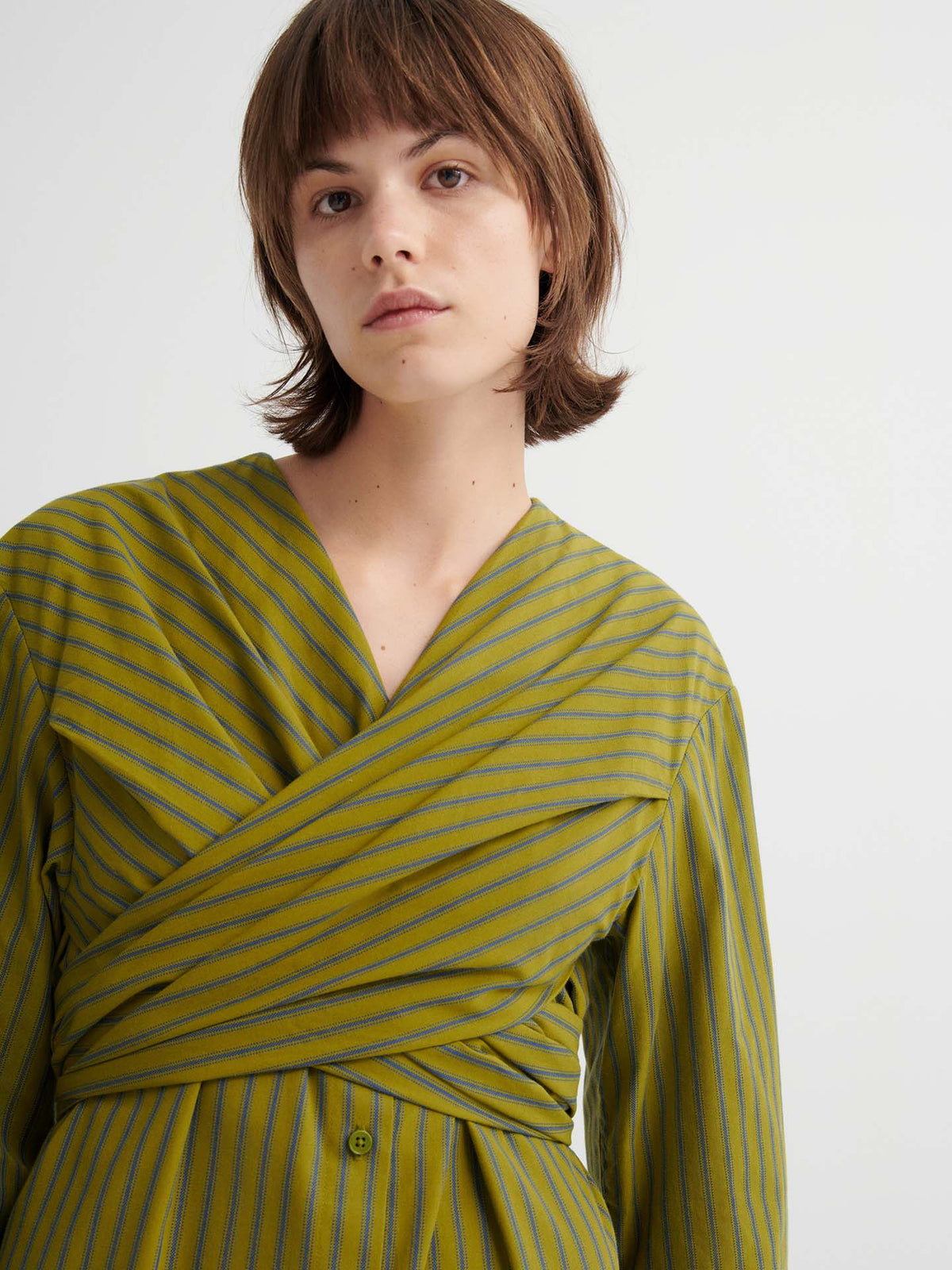 A person with short brown hair wears the Twist Dress – Artisan Stripe, featuring a relaxed fit and thin blue stripes, standing against a plain light background.