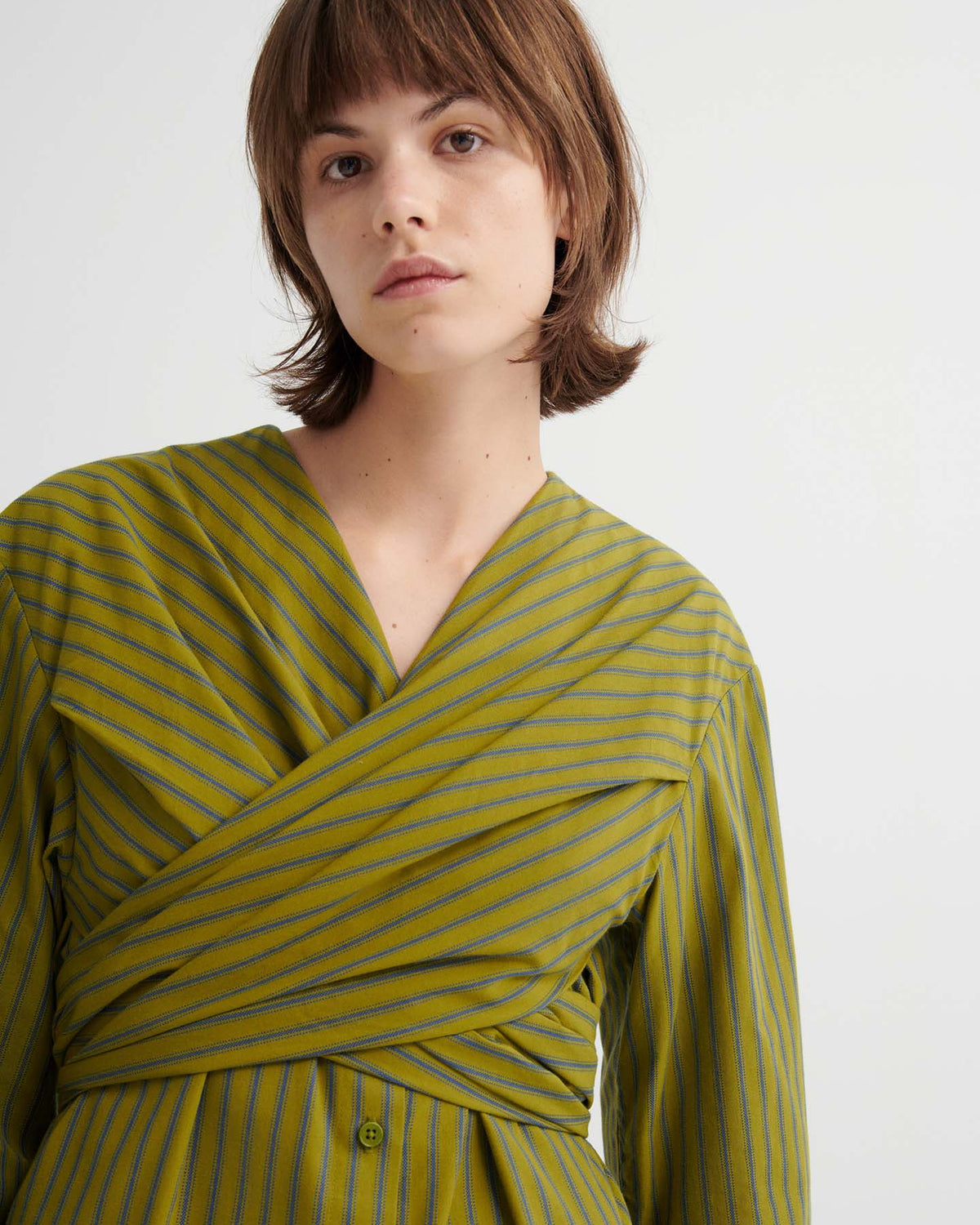 A person with short brown hair wears the Twist Dress – Artisan Stripe, featuring a relaxed fit and thin blue stripes, standing against a plain light background.