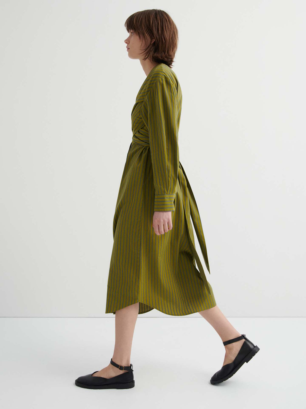 A person with short brown hair walks in profile, wearing the Twist Dress – Artisan Stripe in olive green stripes and black flat shoes against a plain white background.