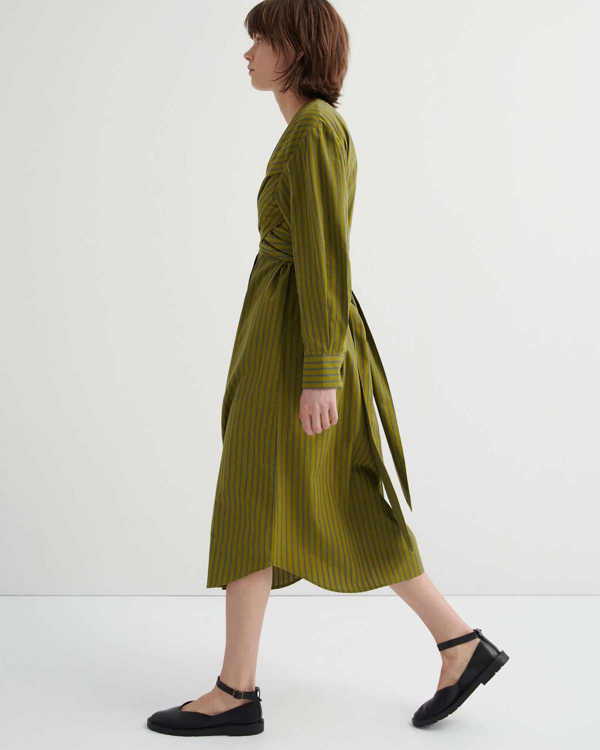 A person with short brown hair walks in profile, wearing the Twist Dress – Artisan Stripe in olive green stripes and black flat shoes against a plain white background.
