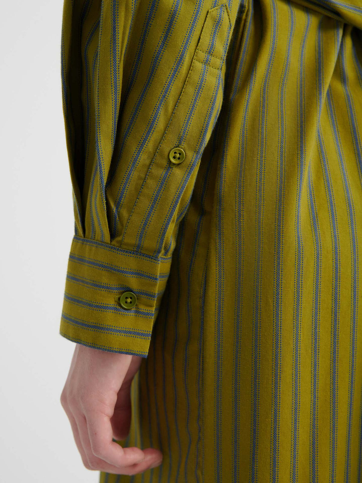 A close-up of a person’s arm wearing the Twist Dress – Artisan Stripe, featuring green and blue stripes with a relaxed fit, highlighting the buttoned cuff on the sleeve and part of their hand.