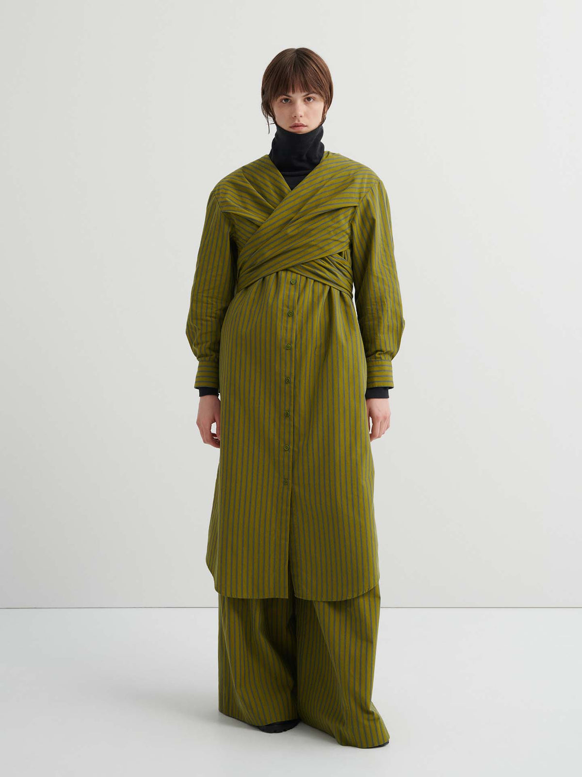 A person stands against a plain background, wearing the relaxed fit Twist Dress – Artisan Stripe over matching wide-leg pants and a black turtleneck.