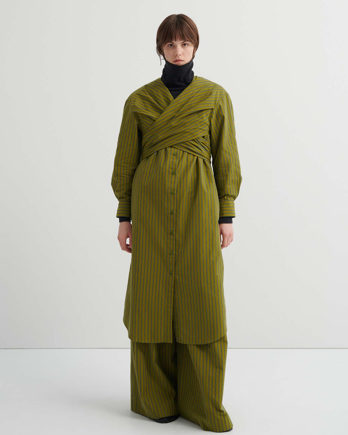 A person stands against a plain background, wearing the relaxed fit Twist Dress – Artisan Stripe over matching wide-leg pants and a black turtleneck.