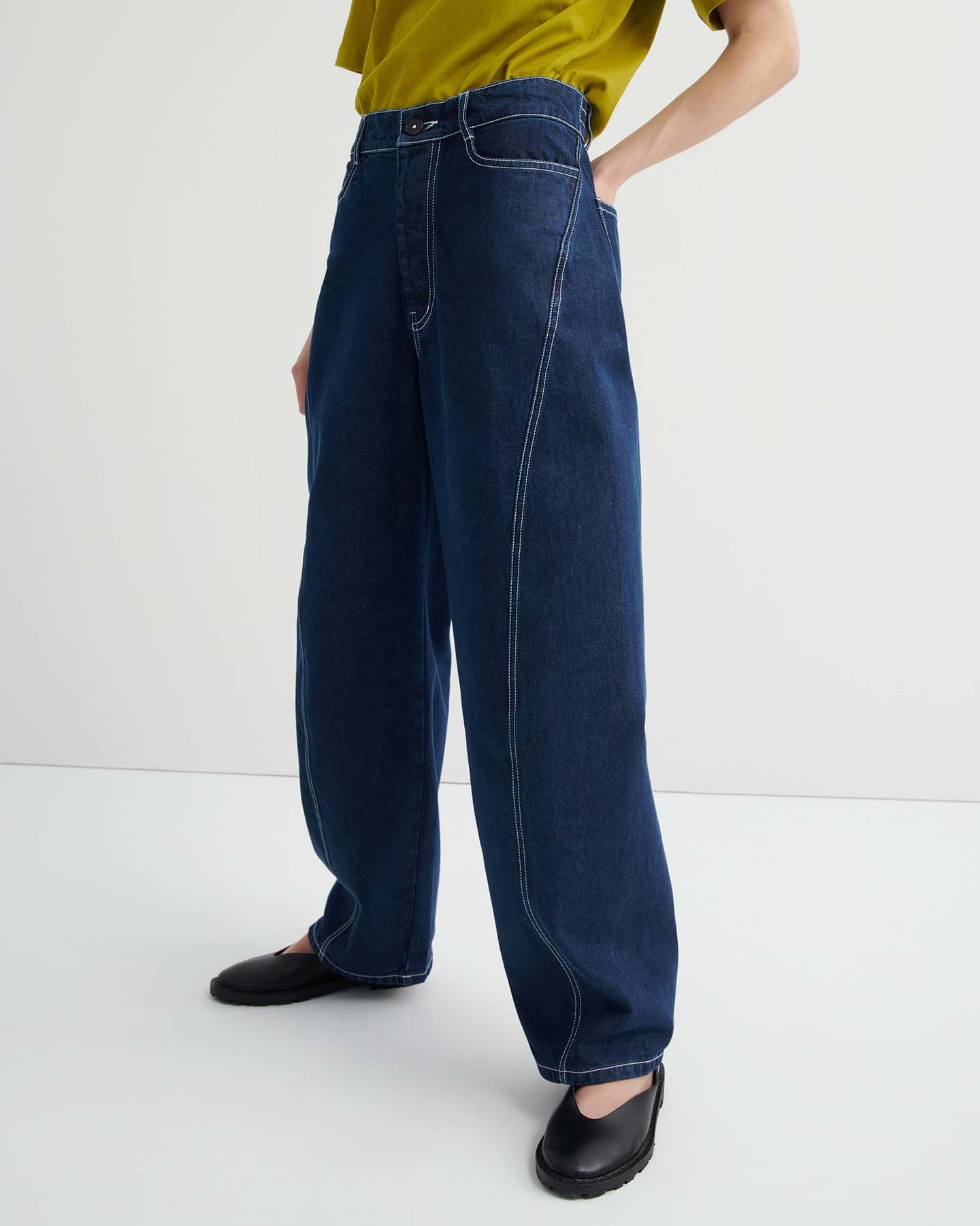 A person stands against a plain background wearing a loose-fitting yellow-green t-shirt, Twist Jeans – Indigo Denim featuring a relaxed, high-waisted fit with white stitching, and black shoes.