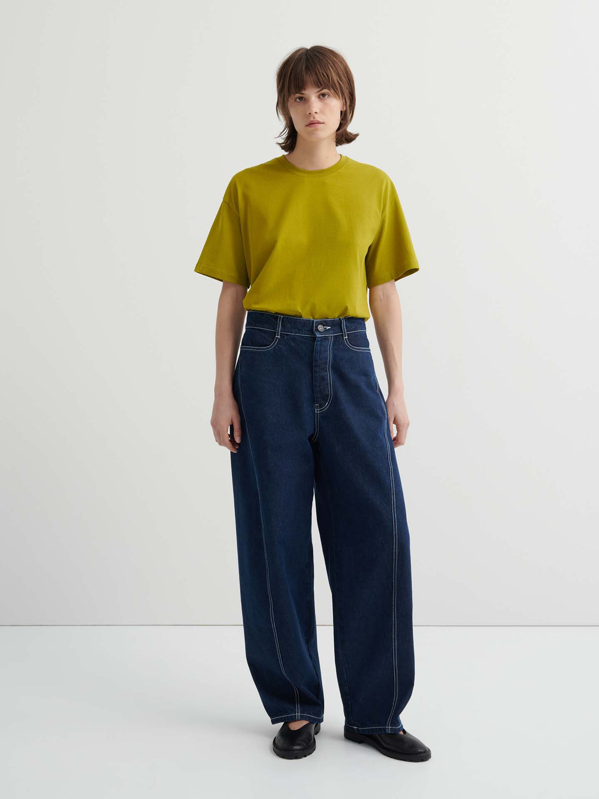 A person stands against a plain background wearing a loose-fitting yellow-green t-shirt, Twist Jeans – Indigo Denim featuring a relaxed, high-waisted fit with white stitching, and black shoes.