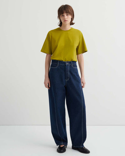 A person stands against a plain background wearing a loose-fitting yellow-green t-shirt, Twist Jeans – Indigo Denim featuring a relaxed, high-waisted fit with white stitching, and black shoes.
