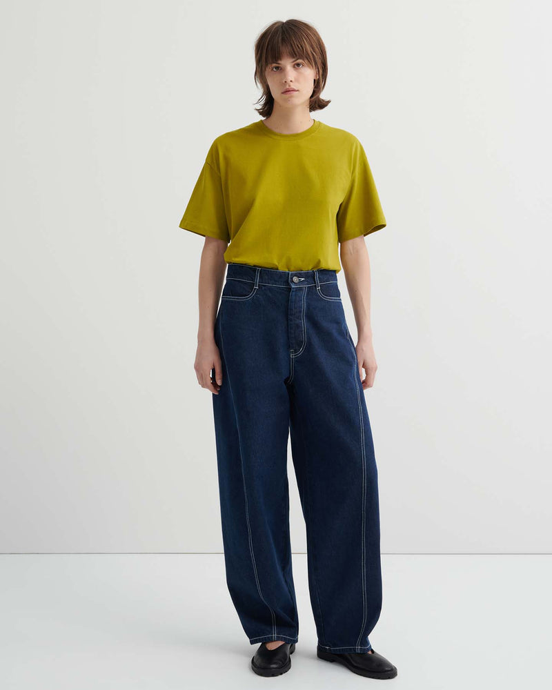 A person stands against a plain background wearing a loose-fitting yellow-green t-shirt, Twist Jeans – Indigo Denim featuring a relaxed, high-waisted fit with white stitching, and black shoes.