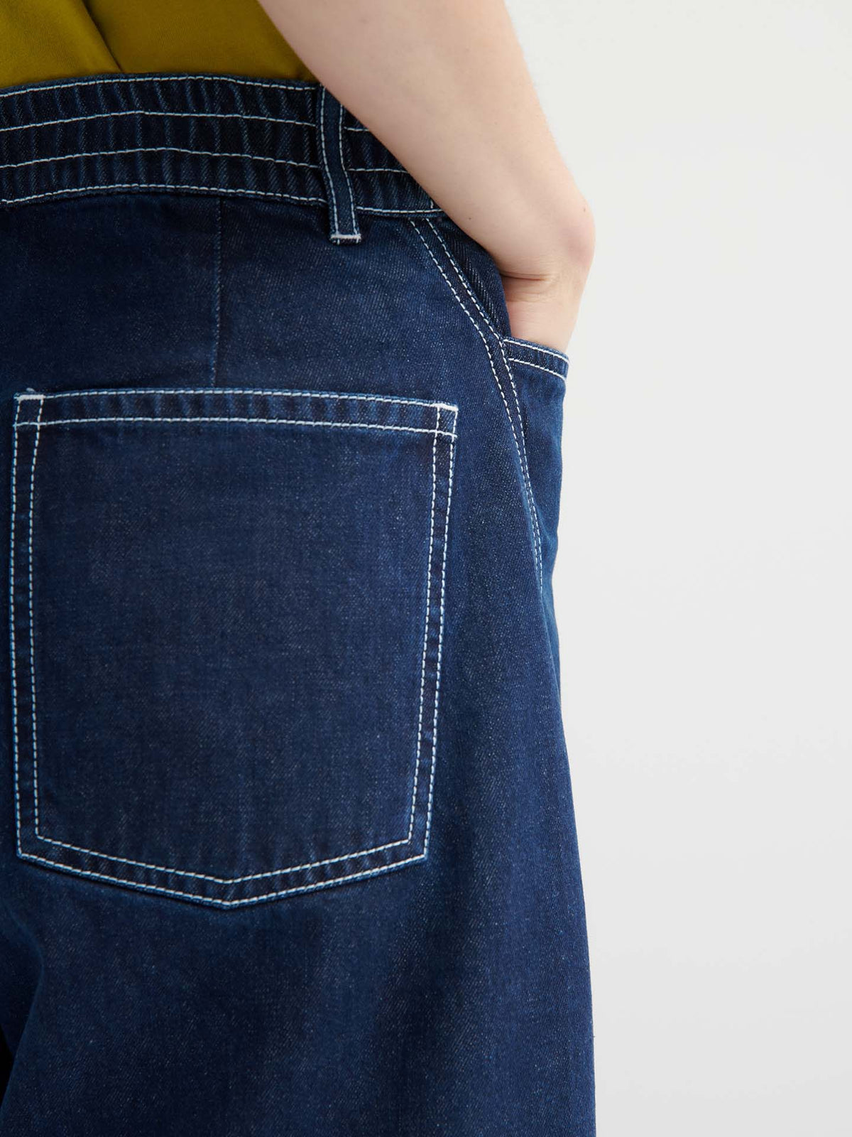 Model wears indigo Twist Jeans with white stitching, styled high-waisted and standing sideways with one hand in their pocket. Green shirt is tucked in.