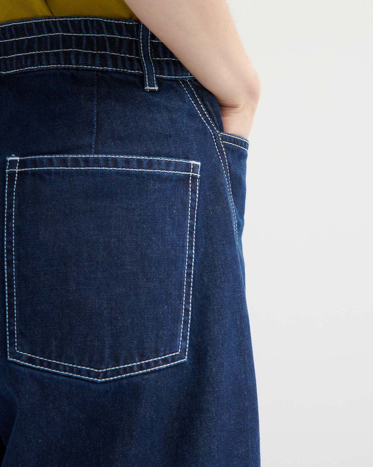 Model wears indigo Twist Jeans with white stitching, styled high-waisted and standing sideways with one hand in their pocket. Green shirt is tucked in.