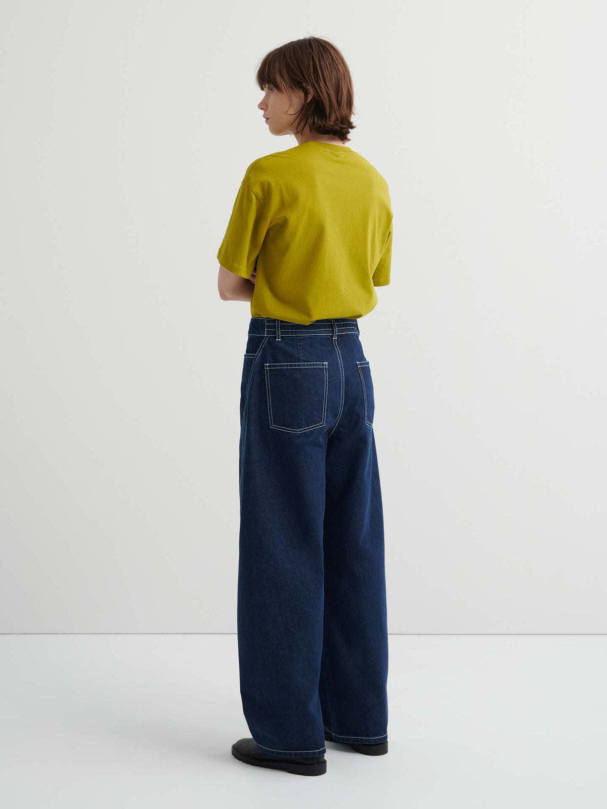 A person stands indoors facing away, wearing a yellow short-sleeve shirt and Twist Jeans – Indigo Denim, featuring a relaxed fit, high waist, and visible back pockets.
