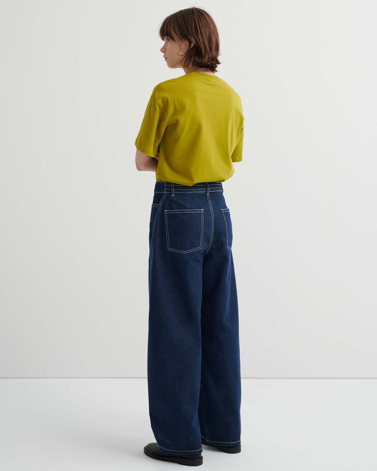 A person stands indoors facing away, wearing a yellow short-sleeve shirt and Twist Jeans – Indigo Denim, featuring a relaxed fit, high waist, and visible back pockets.