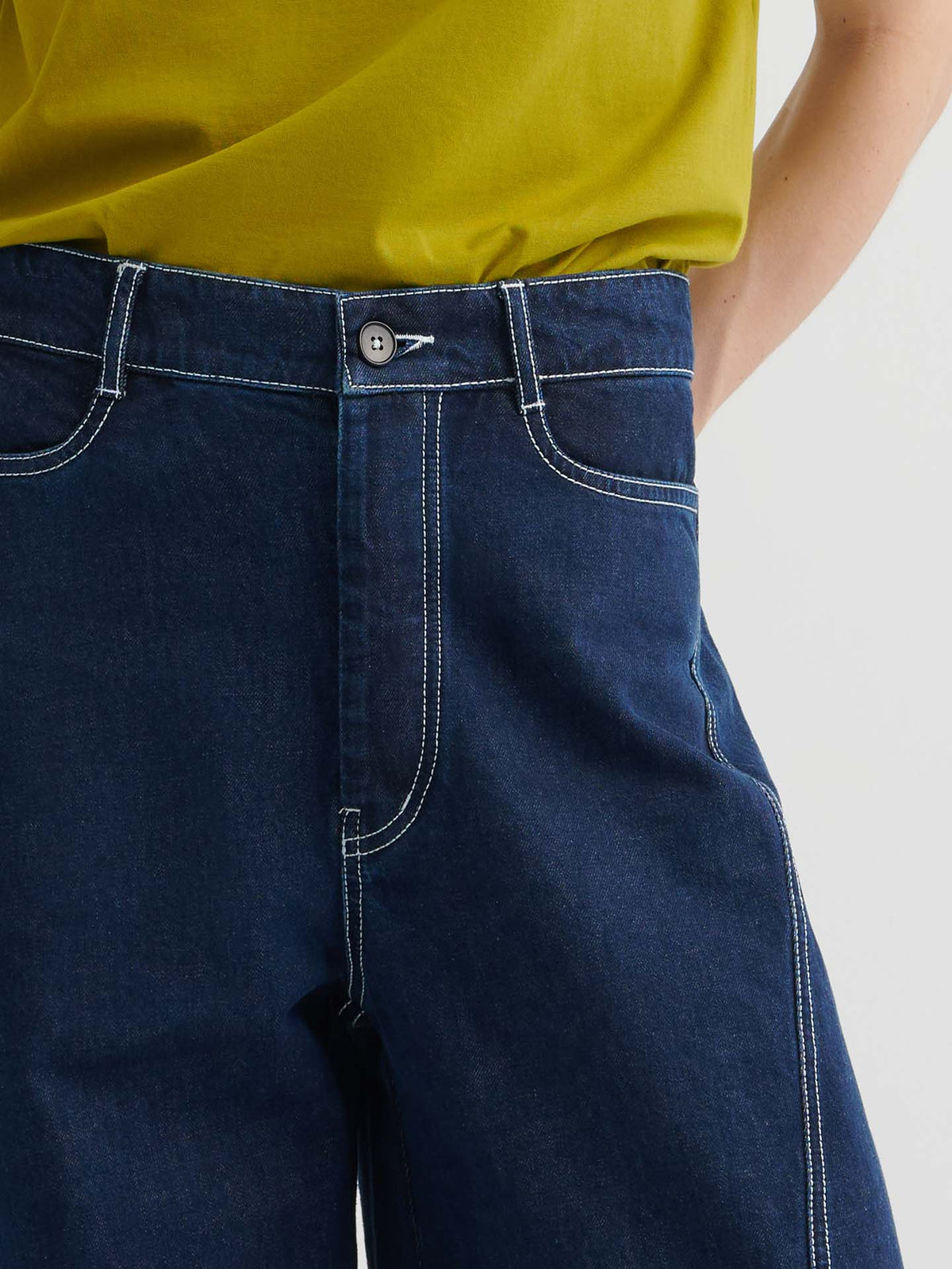 A person wearing Twist Jeans – Indigo Denim, high-waisted with white stitching, paired with a yellow-green top, shown from waist to mid-thigh.