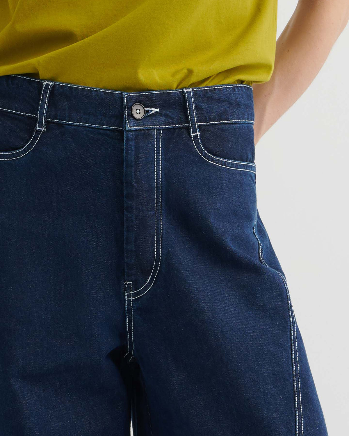 A person wearing Twist Jeans – Indigo Denim, high-waisted with white stitching, paired with a yellow-green top, shown from waist to mid-thigh.